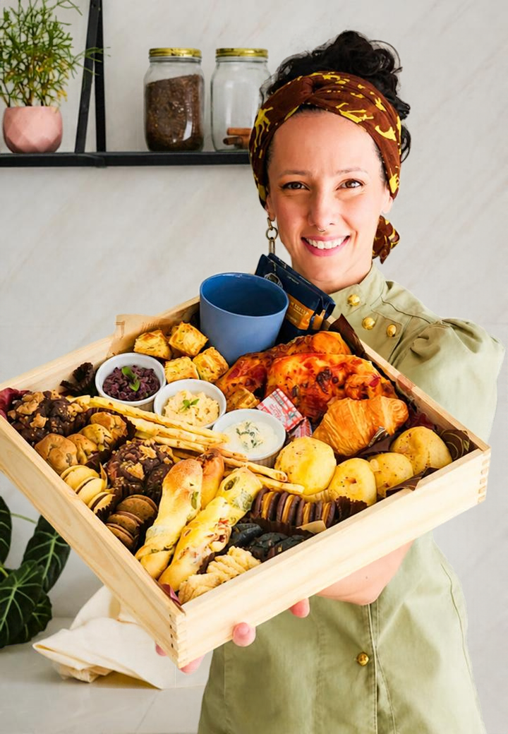 Gio Capostagno sorrindo, vestida de chef e segurando uma caixa de madeira repleta de produtos de bakery